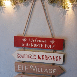 The sign hangs against a backdrop of snow-dusted pine branches and soft twinkling fairy lights, casting warm glimmers across the painted wood. Subtle shadows and muted reds and greens dominate the palette, while a gentle vignette draws focus to the festive signage.
